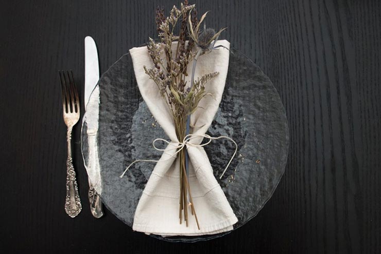 A clear plate on a table with a napkin and dry thistle and lavender flowers tied to it with string, beside a knife and fork.