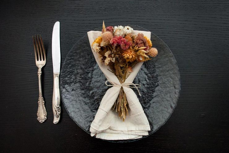 A napkin with a bouquet of dry flowers tied to it on a plate beside a knife and fork.