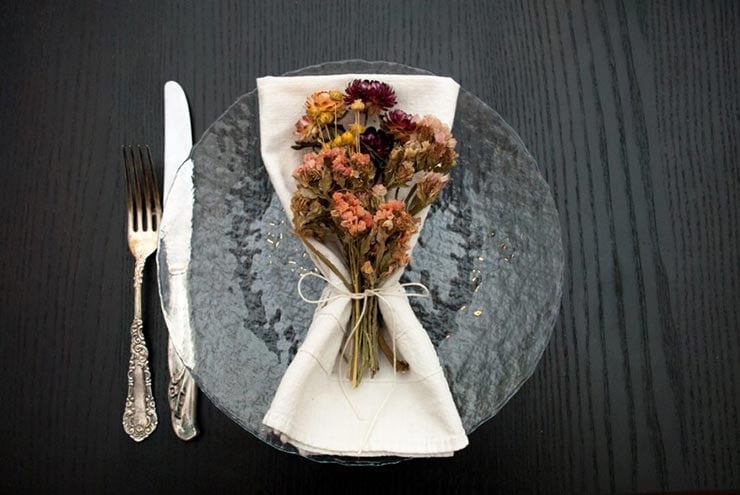 A clear plate on a black table with a napkin and bright and fluffy dry flowers tied to it with string, beside an antique knife and fork.