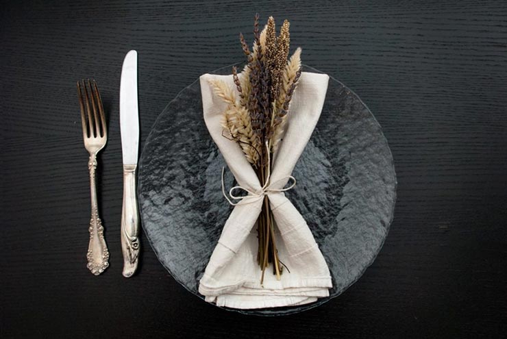 A clear plate on a table with a napkin and dry, purple and white flowers tied to it with string, beside a knife and fork.