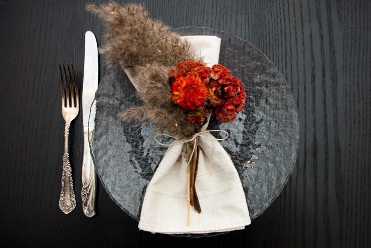 A clear plate on a table with a napkin and bright, fluffy dry flowers tied to it with string, next to a knife and fork.