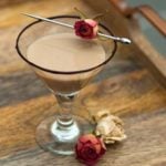A cocktail on a wooden tray, garnished with a chocolate rim and a small rose, with 2 roses at its base.