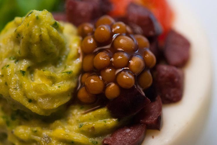 A deviled egg on a marble plate, garnished with mustard caviar, diced olives, a cherry tomato and small leaf of basil.