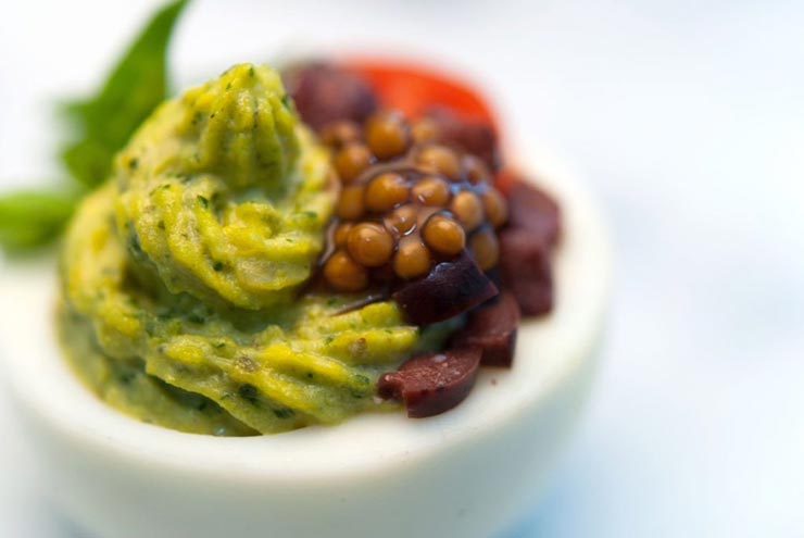 A green deviled egg, garnished with mustard caviar, diced olives, a tiny sliced tomato and a small leaf of basil.