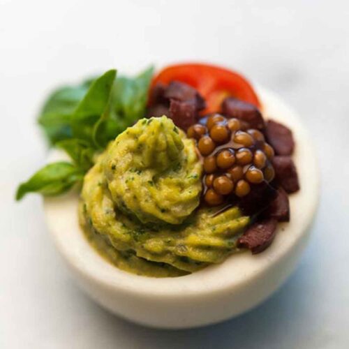 A green deviled egg, garnished with mustard caviar, diced olives, a tiny sliced tomato and a small leaf of basil.