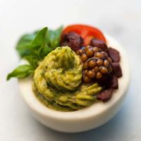 A green deviled egg, garnished with mustard caviar, diced olives, basil leaves and a cherry tomato slice.