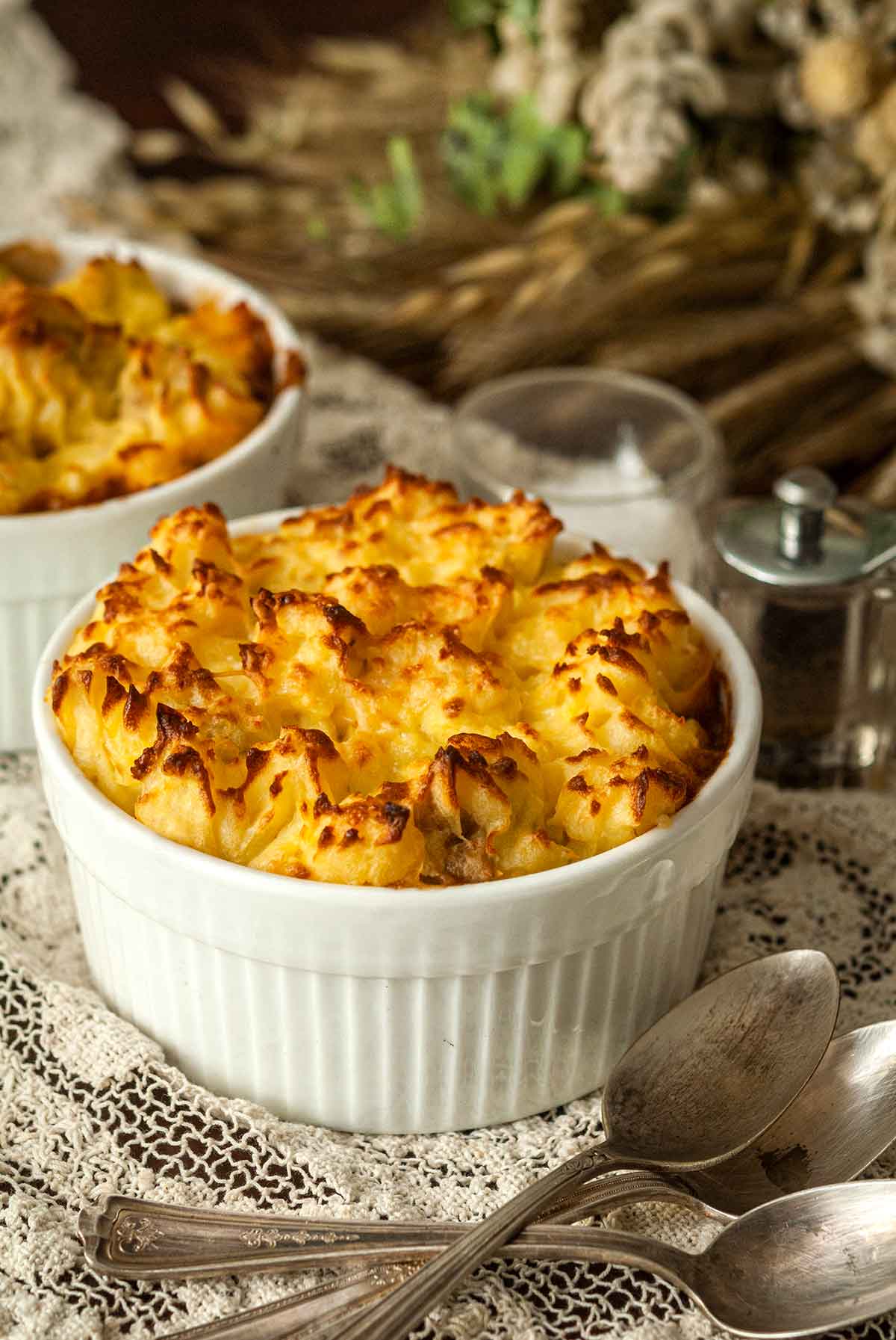 2 shepherd's pies in bowls with crispy tops on a lace table cloth beside spoons.