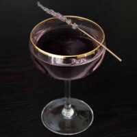 A purple cocktail on a black table, garnished with a sprig of lavender.