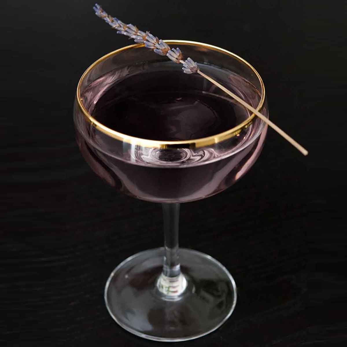 A purple cocktail on a black table, garnished with a sprig of lavender.