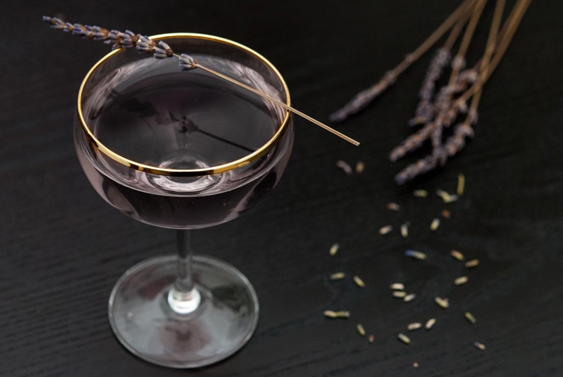 A purple cocktail on a black table, garnished with a sprig of lavender.
