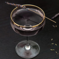 A cocktail on a dark table, garnished with a sprig of lavender, with a few scattered lavender flowers on the table.