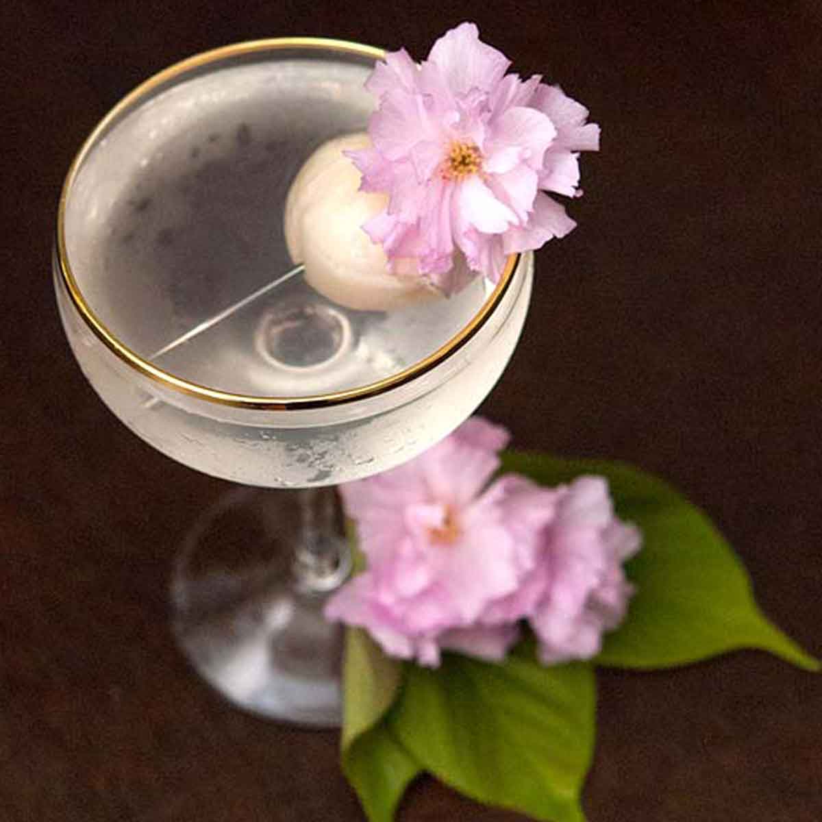 A cocktail garnished with a lychee and cherry blossom flowers, with flowers and a few leaves at its base.