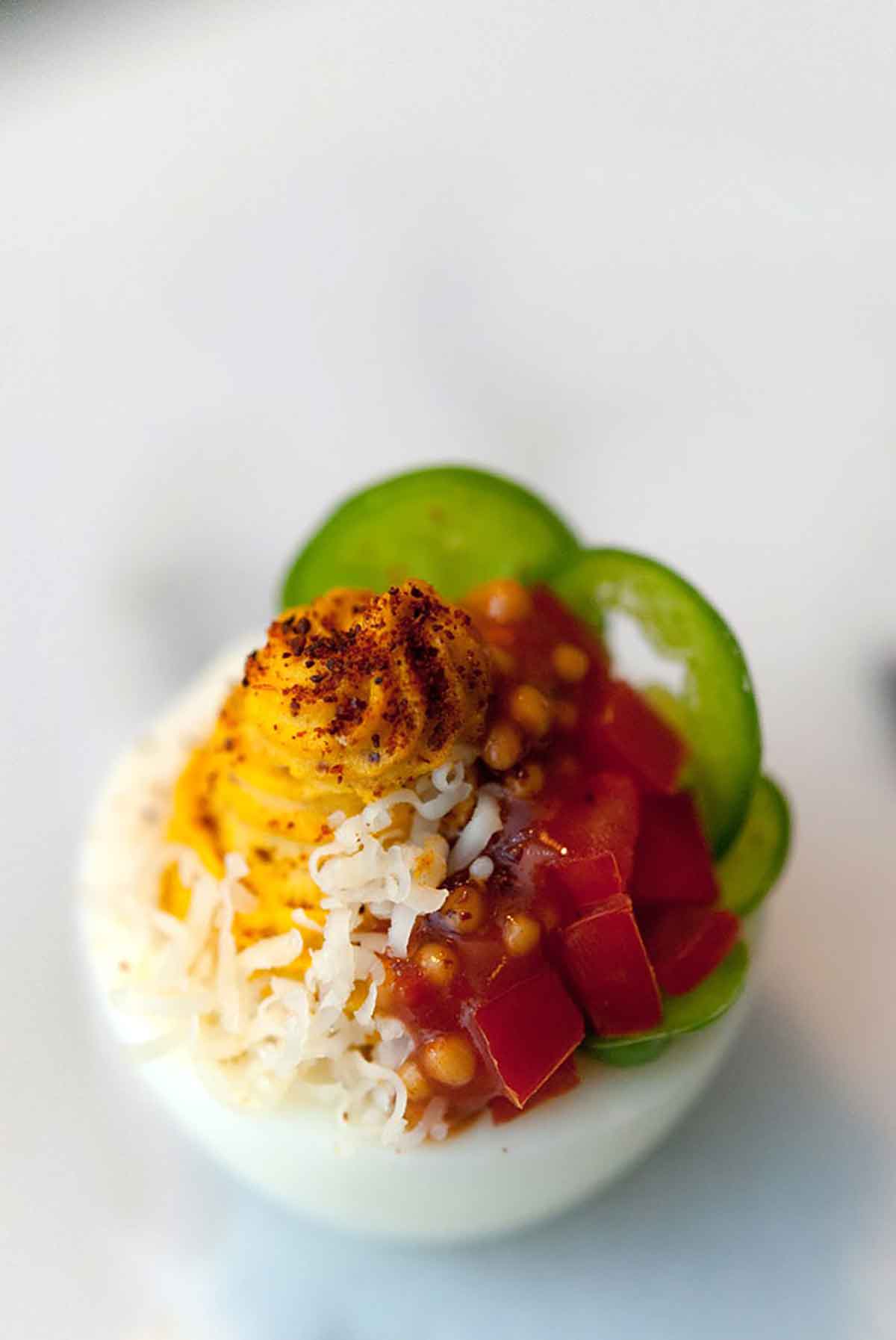 A deviled egg, garnished with sliced jalapeños, diced tomatoes, mustard caviar, shredded cheese and paprika.