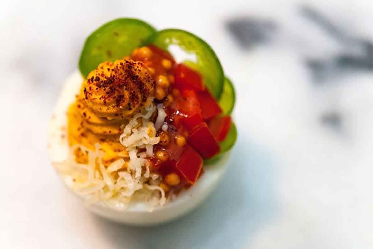 A deviled egg, garnished with sliced jalapeños, diced tomatoes, mustard caviar, shredded cheese and paprika.