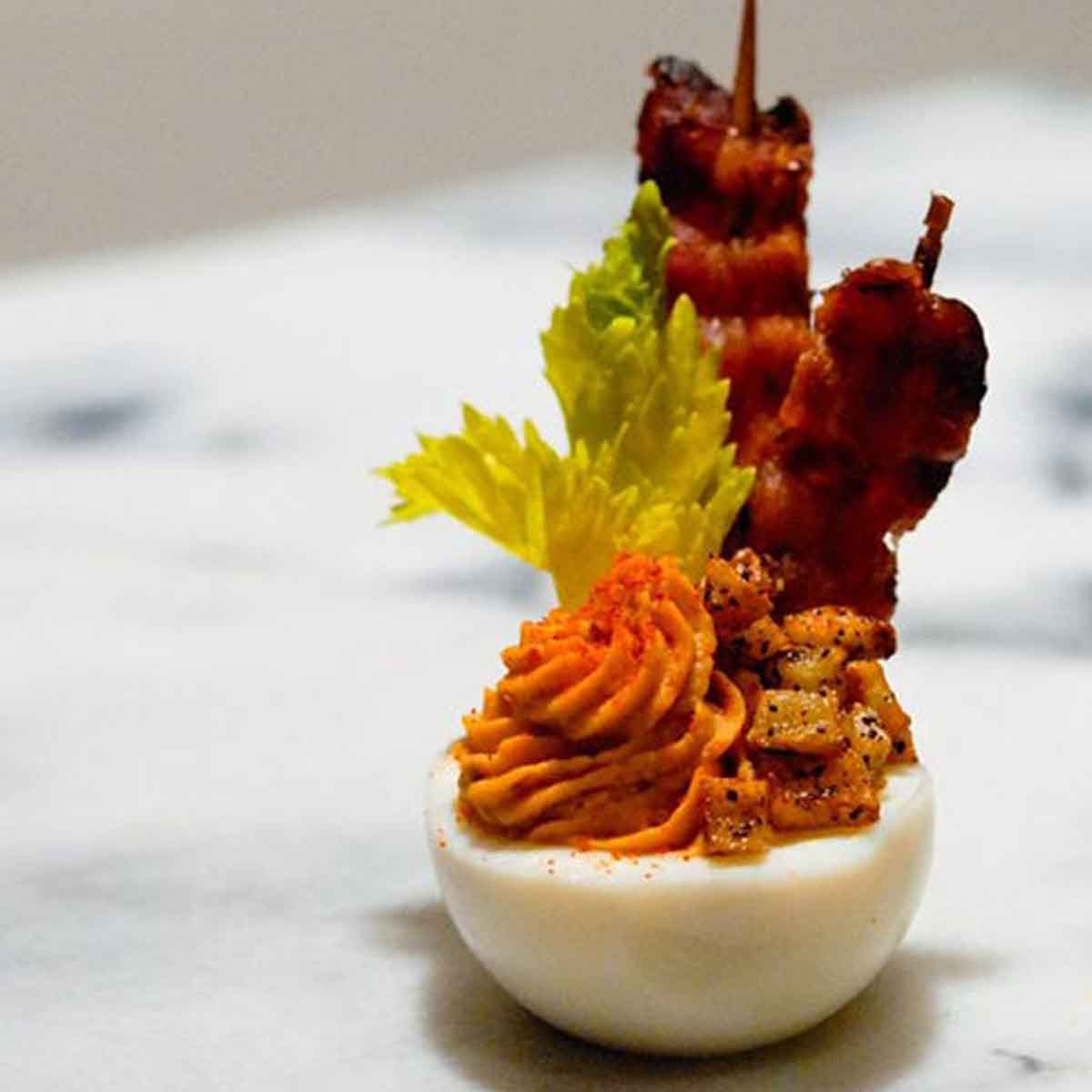 A deviled egg with bacon, celery, and tiny hash browns on a marble table.