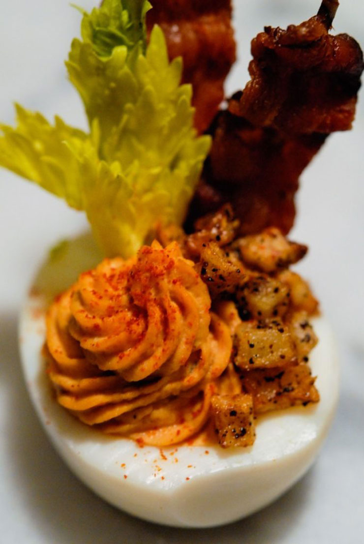 A closeup of a deviled egg with bacon, celery, and tiny hash browns on a marble table.