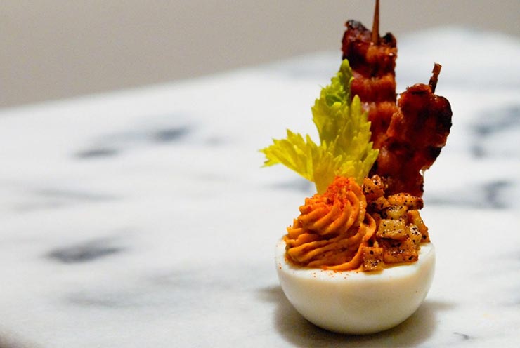 A deviled egg with bacon, celery, and tiny hash browns on a marble table.