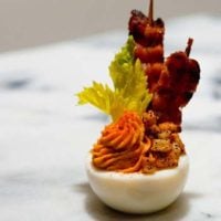 A deviled egg on a marble plate, garnished with small hash browns, a celery leaf and bacon strips.