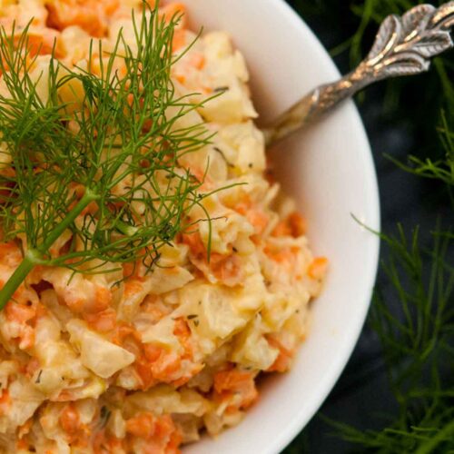 A bowl of fennel & carrot coleslaw in a small white bowl, garnished with a fennel frond next to larger fronds on a table.