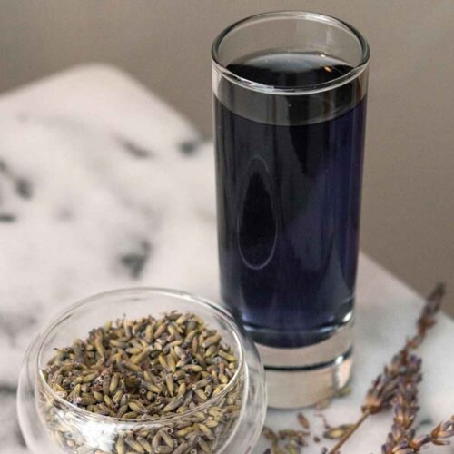 A small cup of cocktail lavender next to a small bowl of dry lavender flowers on a marble table.