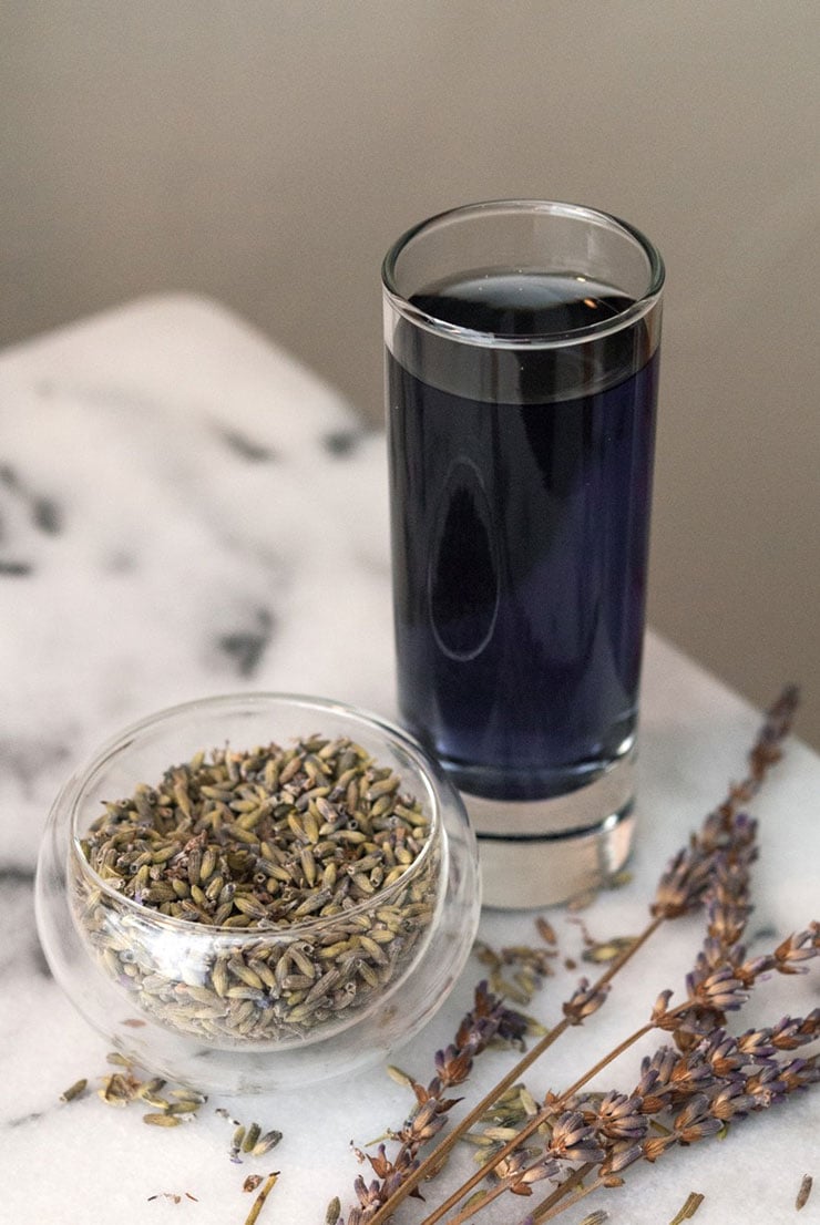 A small cup of cocktail lavender next to a small bowl of dry lavender flowers on a marble table.