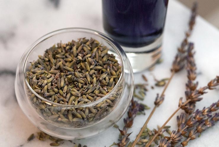 A small bowl of lavender on a marble plate beside a glass of lavender liquor, and a few scattered flowers beside it.