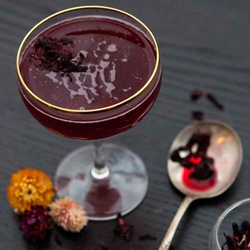 A cocktail on a table with 3 small flowers at its base and a spoon, wet with hibiscus tea, and sprinkled with flowers.