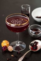 A cocktail on a table with 3 small flowers at its base and a spoon, wet with hibiscus tea, and sprinkled with flowers.