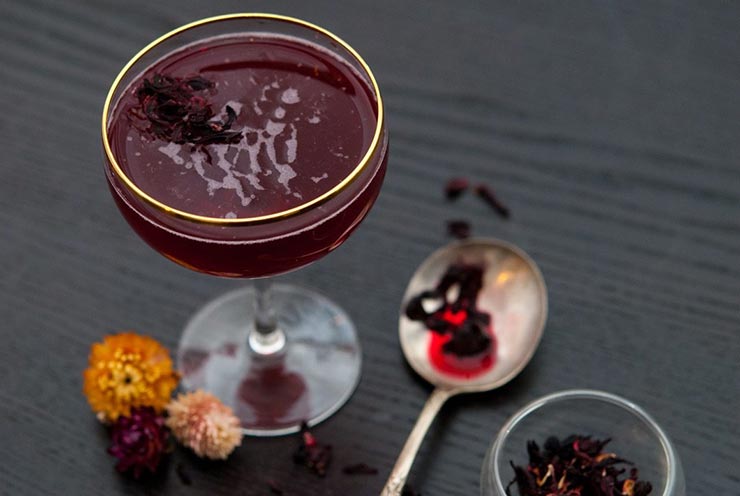 A cocktail on a table with 3 small flowers at its base and a spoon, wet with hibiscus tea, and sprinkled with flowers.