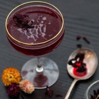 A cocktail on a table with 3 small flowers at its base and a spoon, wet with hibiscus tea, and sprinkled with flowers.