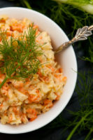 A bowl of fennel & carrot coleslaw in a small white bowl, garnished with a fennel frond next to larger fronds on a table.