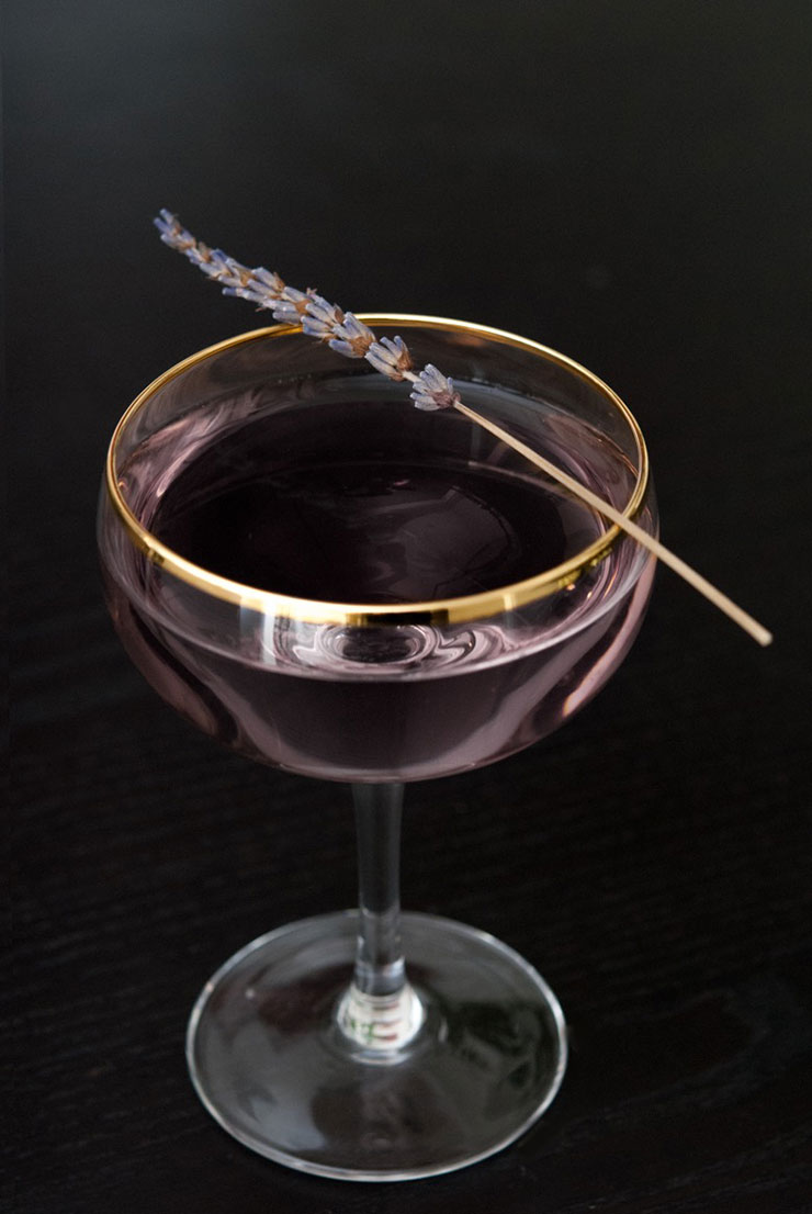 A purple cocktail on a black table, garnished with a sprig of lavender.