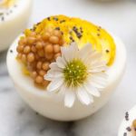 A deviled egg on a marble plate, garnished with poppy seeds, mustard caviar and a small daisy.