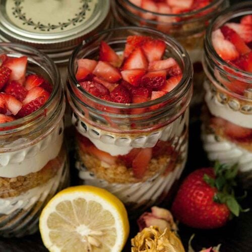 6 jars of mousse with strawberry and cookie crumbles on a table, surrounded by flowers, a strawberry and a lemon slice.