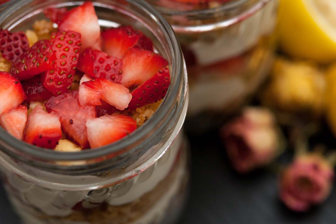 A jar full of strawberries on top of mousse with dry flowers and lemons in the background on a black table.