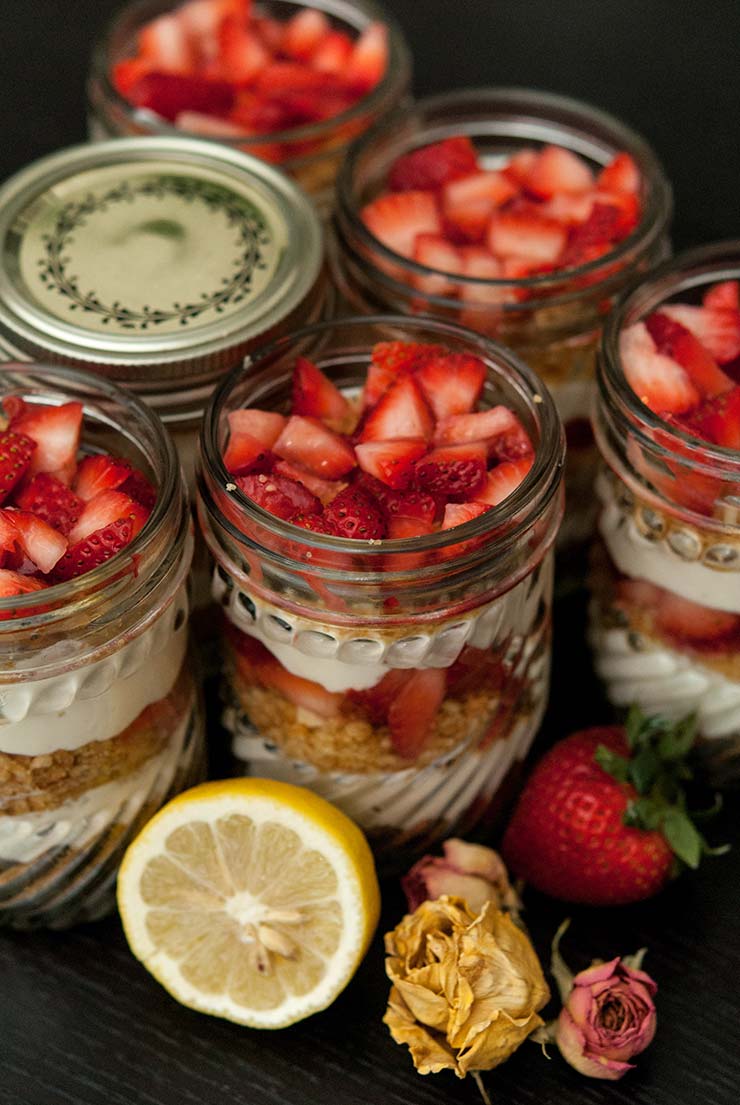 6 jars of mousse with strawberry and cookie crumbles on a table, surrounded by flowers, a strawberry and a lemon slice.