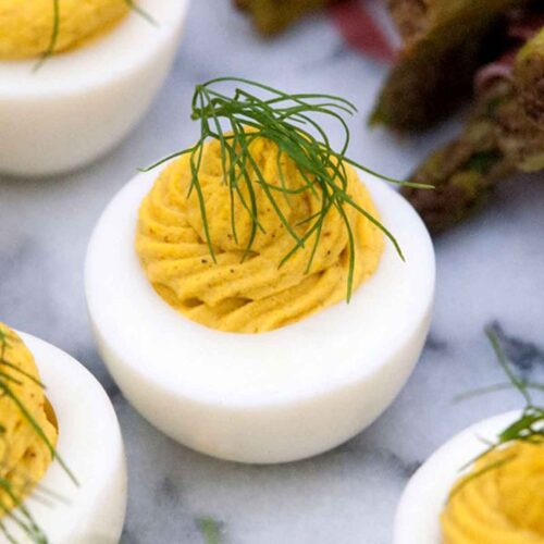 3 deviled eggs on a marble slate, garnished with a sprig of fresh dill, next to prosciutto-wrapped asparagus.