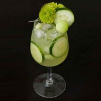 A green cocktail on a black table, garnished with aren flowers, cucumber and a ball of honeydew with cucumbers in the glass.