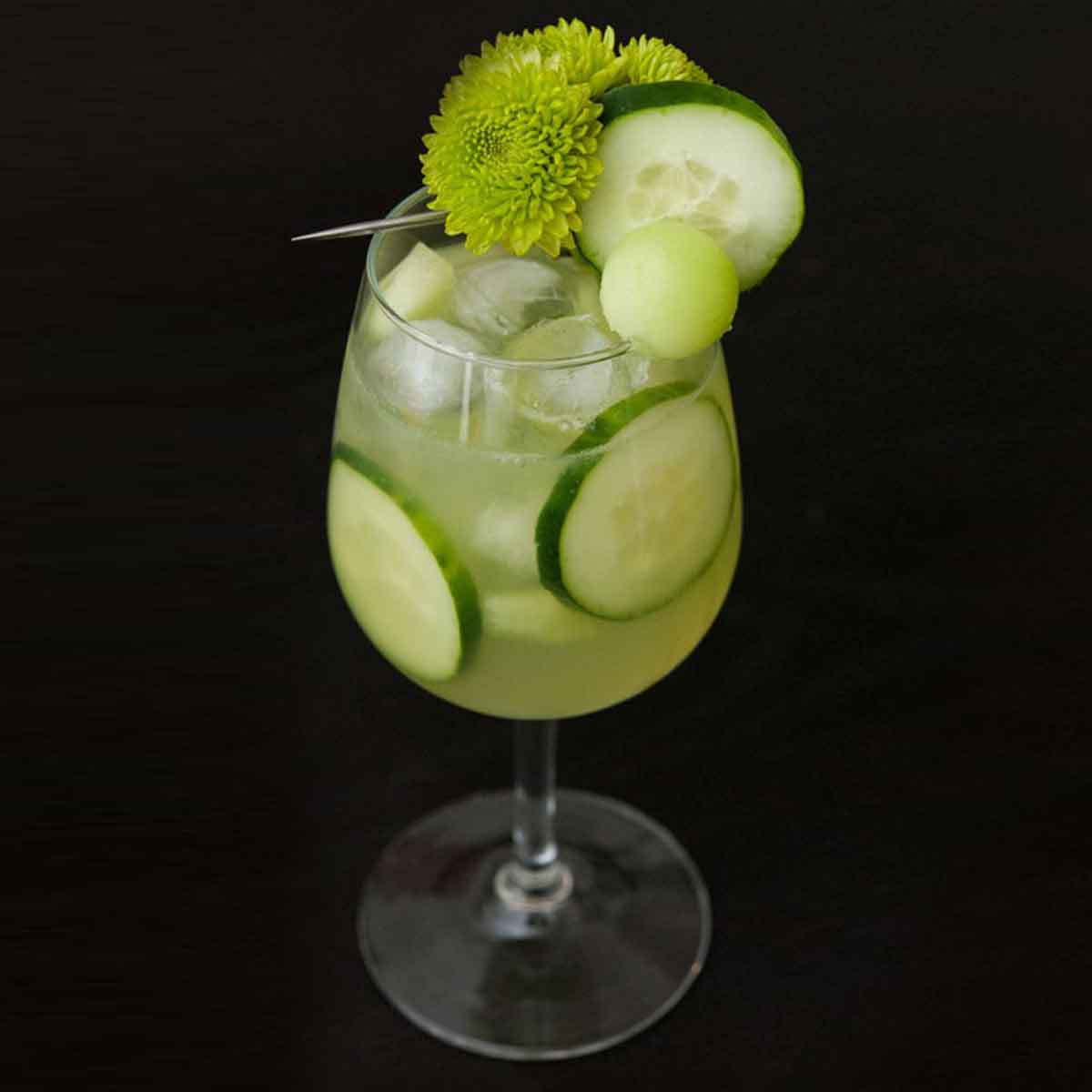 A green cocktail on a black table, garnished with aren flowers, cucumber and a ball of honeydew with cucumbers in the glass.