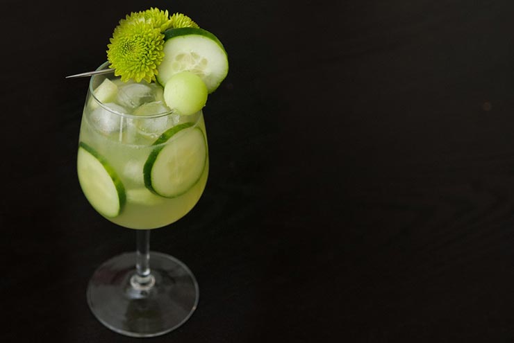 A cocktail in a glass with ice, cucumber slices and garnished with green flowers, cucumber and a honeydew ball.