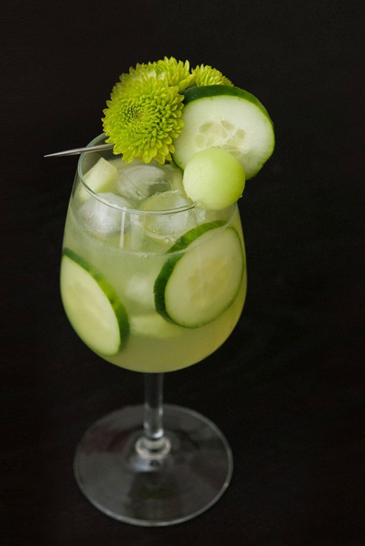 A cocktail in a glass with ice, cucumber slices and garnished with green flowers, cucumber and a honeydew ball.
