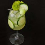 A cocktail in a glass with ice, cucumber slices and garnished with green flowers, cucumber and a honeydew ball.