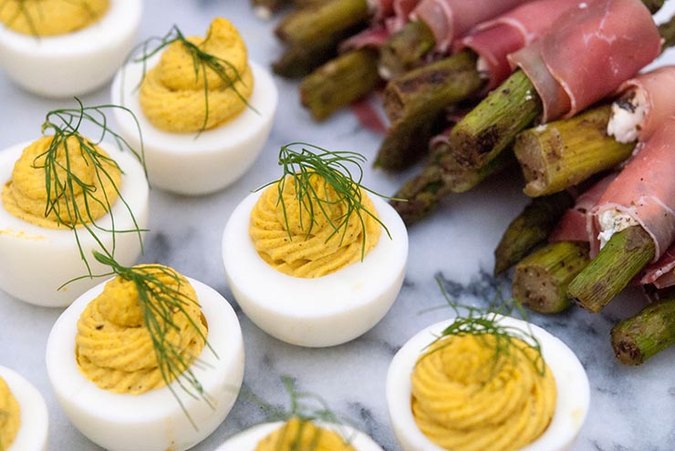 7 deviled eggs on a marble slate, garnished with a sprig of fresh dill, next to prosciutto-wrapped asparagus.