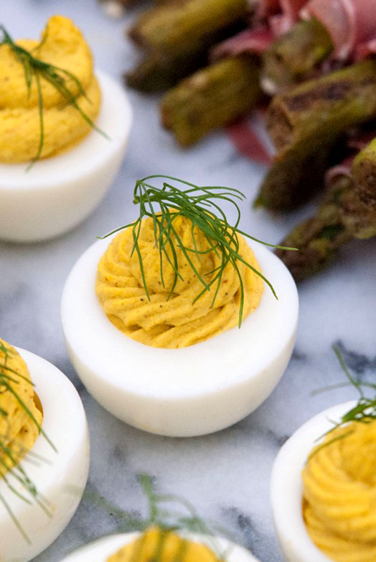 3 deviled eggs on a marble slate, garnished with a sprig of fresh dill, next to prosciutto-wrapped asparagus.