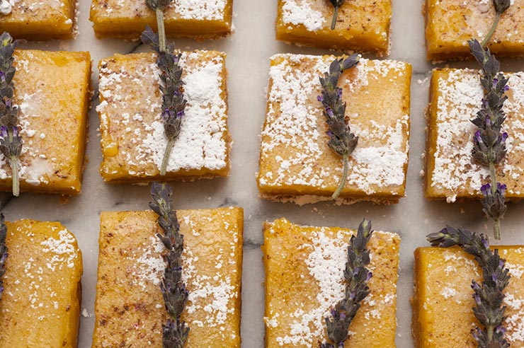 12 lavender lemon bars on a white marble table garnished with fresh lavender and powdered sugar.