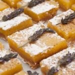 6 lavender lemon bars on a marble plate, topped with sprigs of lavender and sprinkled with powdered sugar.