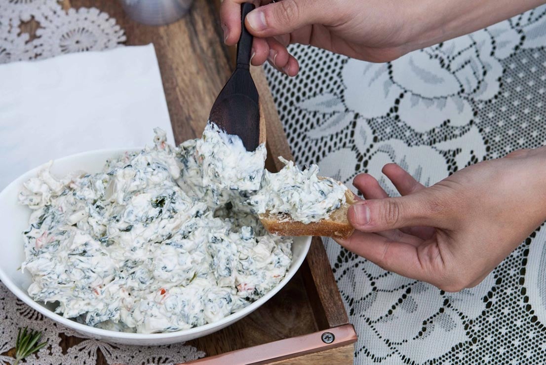 A hand scooping spinach dip onto bread.