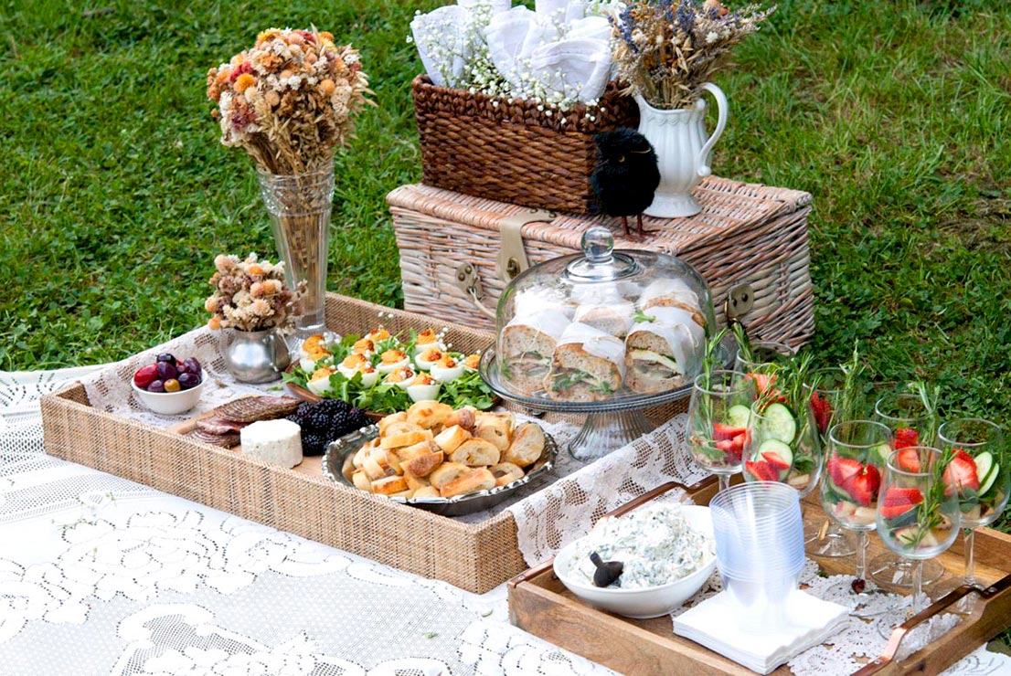 An elaborate picnic spread. Trays of food, flowers, picnic basket and glassware on a blanket.