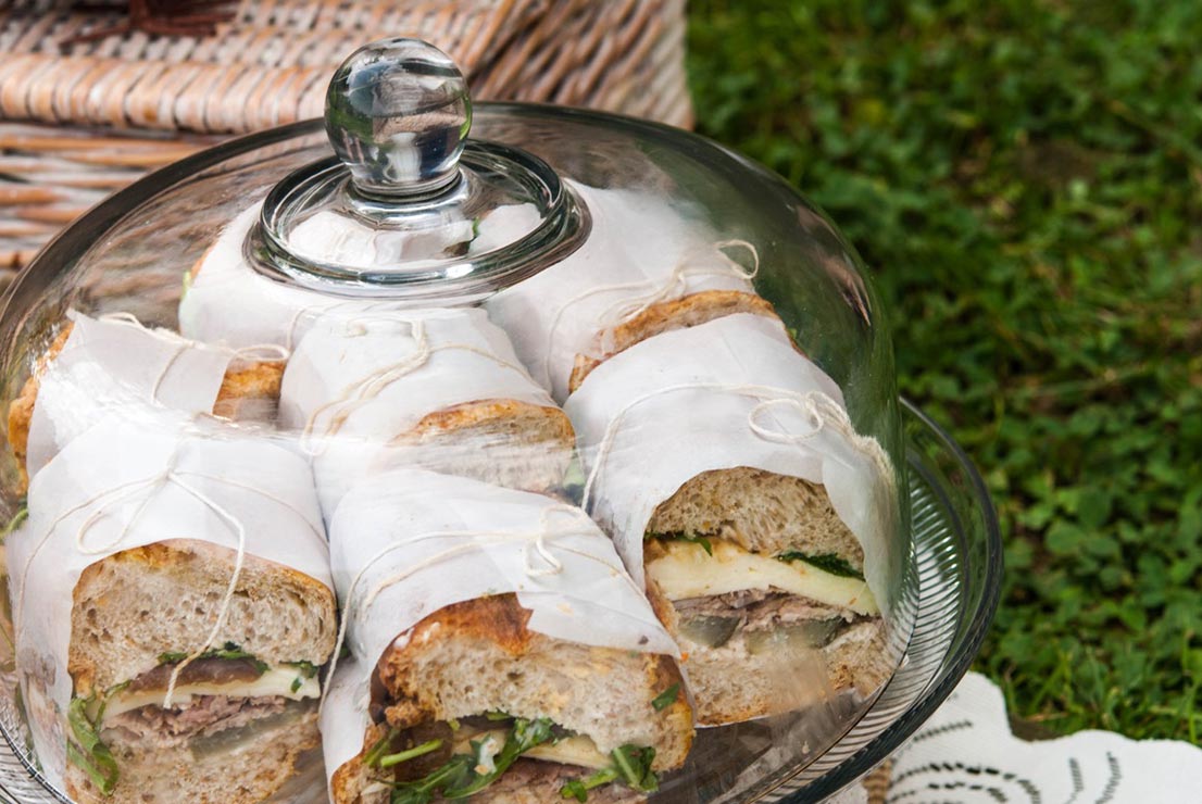 Sandwiches wrapped in paper, tied with string on a cake platter with grass and a picnic basket in the background.