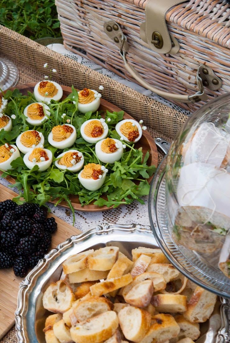 A picnic spread of baguette, a plate of deviled eggs and sandwiches beside a picnic basket.